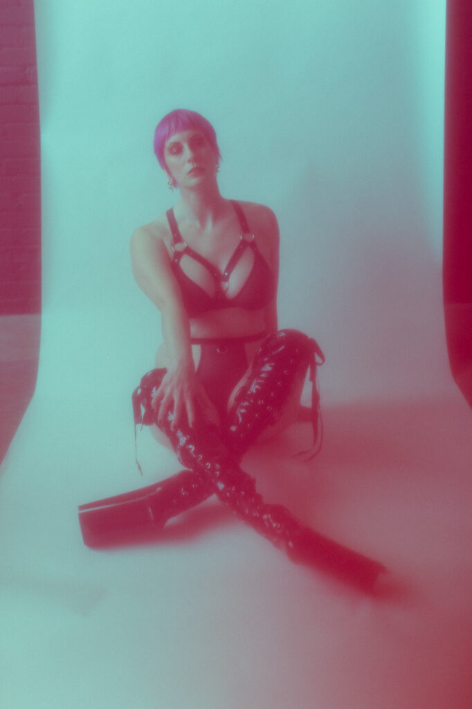 Self portrait with a rose tint with a white back drop dressed in black harnesses and tall boots.